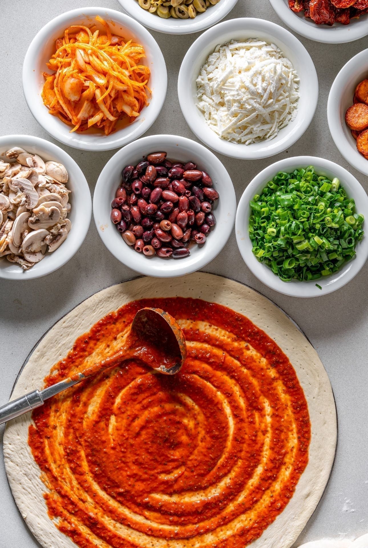 Pizza dough with tomato sauce and various toppings in bowls: mushrooms, olives, cheese, onions, pepperoni.